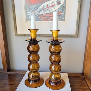 May include: Two amber-colored glass candle holders with white candles. Each holder has a round base, a series of stacked spheres, and a flared top. The candle holders are on a white book, with a framed fish print in the background.