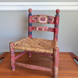 May include: A small wooden chair with a woven rush seat and a distressed red painted finish. The backrest features two horizontal slats decorated with hand-painted floral motifs in white, green, and yellow, accented by dark blue turned finials.