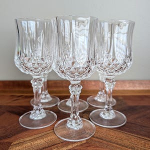May include: A set of five clear glass goblets featuring a diamond-cut pattern on the lower bowl and a faceted stem. Each goblet has a smooth, flared rim and a circular base, reflecting light through the intricate, textured glass design.