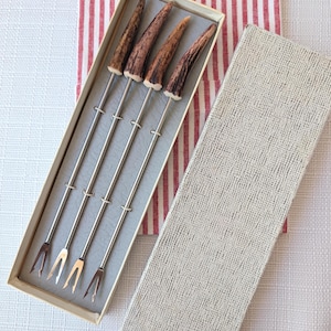 May include: A set of four silver-toned metal appetizer forks with textured brown antler-style handles, presented in a rectangular beige box with a matching lid. The forks feature two-pronged tips and are arranged side-by-side within the box.