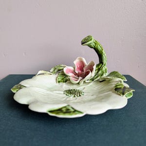 May include: A ceramic floral dish with a light green base shaped like a flower. The dish features a pink and white flower in the center, with green leaves and a curved stem. The dish is on a dark blue surface.