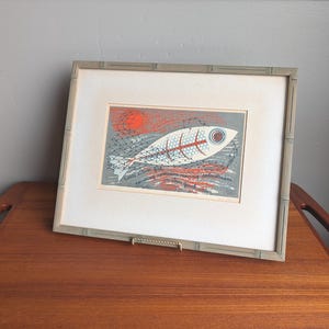 May include: Framed art print featuring a stylized white fish with red and blue accents, set against a gray background with a red sun. The print is in a bamboo-style frame, displayed on a wooden surface.