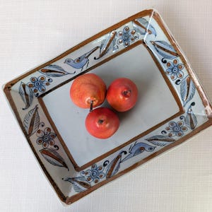 May include: A rectangular ceramic tray with a white center and a decorative border featuring hand-painted blue birds, brown leaves, and blue flowers. Three small, realistic red pears are placed in the center of the tray.
