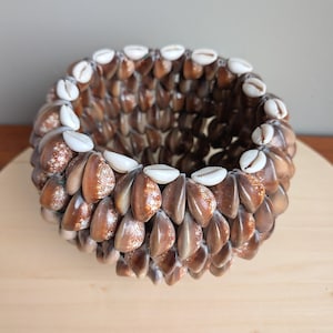 May include: A round decorative bowl crafted from numerous brown and white seashells. The bowl's exterior is composed of tightly arranged, glossy brown shells, while the rim features a contrasting row of white shells. The bowl sits on a light-colored wooden surface.
