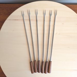 May include: Six vintage fondue forks with stainless steel tines and long, slender stems. The handles are made of dark brown wood. The forks are arranged on a light-colored wooden surface.