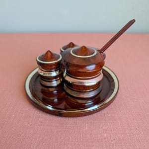 May include: A condiment set featuring a circular tray, two small shakers, and a lidded jar with a matching spoon. Each piece is crafted from a dark brown, polished material with a wood-grain appearance, accented by thin, bright metallic silver bands.