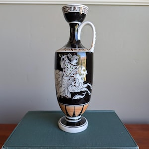May include: Black ceramic vase with a white handle and a wide base. The vase features a Greek mythological scene in white against a black background. The vase is approximately 10 inches tall.