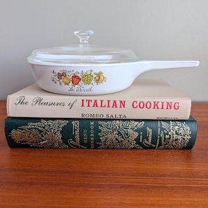 May include: White, vintage cookware with a clear glass lid and a long handle. The pan features a colorful fruit and vegetable design and the text "Le Persil". It rests on top of two cookbooks.