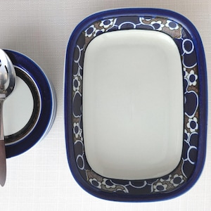 May include: A rectangular ceramic dish with a white center and a wide blue border featuring a dark blue and white floral pattern, accompanied by a matching round bowl and a silver-toned metal spoon with a dark handle positioned to the left.