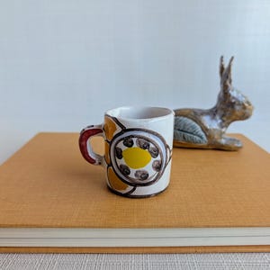 May include: A small ceramic mug with a white base featuring a hand-painted floral design in yellow, brown, and red. The mug has a curved handle and a cylindrical shape. Beside the mug sits a small, brown, textured ceramic rabbit figurine.