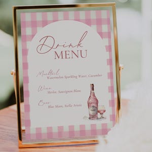 Op de afbeelding: Een roze en wit geruit bord met een gouden lijst, met een drankenmenu met de titel "Drink Menu". Het menu bevat een mocktail, wijn en bier. De mocktail is watermeloen-sprankelend water met komkommer. De wijnen zijn Merlot en Sauvignon Blanc. De bieren zijn Blue Moon en Stella Artois.