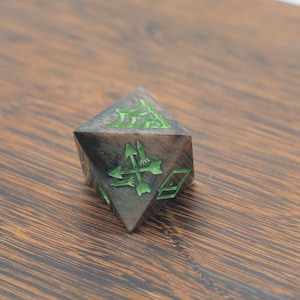 May include: A multi-sided, dark-colored die with green symbols. The die has a geometric shape and is resting on a wooden surface. The green symbols include arrows and other geometric shapes.