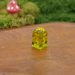 May include: A translucent lime green four-sided die with a rounded top and flat base. The die features engraved numbers on its sides, including a clearly visible number four and number three, filled with a contrasting yellow pigment.