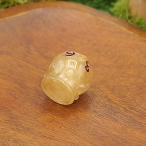 Peut inclure: Un petit dé à coudre doré avec des marques violet foncé. Le dé à coudre est posé sur une surface en bois, mettant en valeur son design unique. Cet objet de collection est un article décoratif.