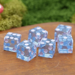 May include: A set of six translucent dice made of clear resin, each featuring embedded circular pips in alternating shades of light blue and soft lavender. The dice are arranged in a cluster, showcasing their cubic shape and smooth, polished surfaces.