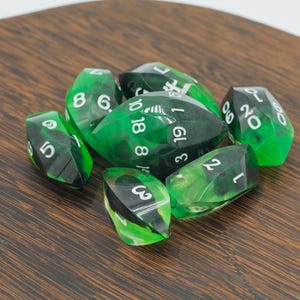 May include: A collection of seven polyhedral dice with a green and black marbled design. Each die has white numbering. The dice are arranged on a dark brown wooden surface.