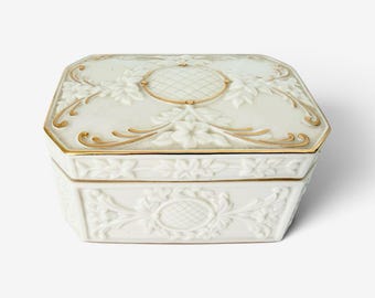 May include: A rectangular white porcelain trinket box featuring embossed floral and leaf patterns on the lid and sides. The design includes a central lattice motif on the lid and gold-painted trim along the edges and decorative elements.