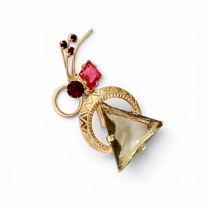 Peut inclure: Une broche dorée ornée d'une grande pierre triangulaire brun clair fixée dans un cadre circulaire texturé. Le design comprend une petite pierre rouge en forme de losange, une pierre rouge foncé ronde et trois minuscules accents rouges.