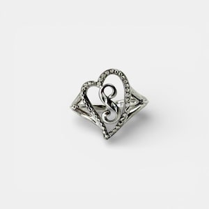 May include: A silver ring with a heart-shaped design and a decorative swirl in the centre. The heart is outlined with small, round embellishments. The ring has a delicate, openwork design.