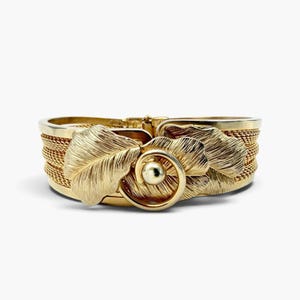 May include: A gold-tone cuff bracelet featuring a wide, woven mesh band. The center is adorned with two textured, leaf-shaped metal elements and a central circular motif with a smooth, polished gold sphere at its core, creating a layered design.