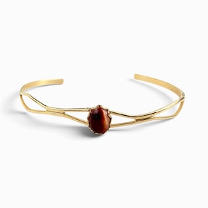 May include: A gold-toned open cuff bracelet featuring a split-shank design that converges at the center. The focal point is a single, oval-shaped, deep reddish-brown stone with a smooth, polished surface set within a simple gold-colored metal bezel.