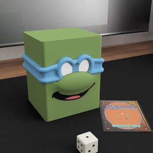 May include: A green, cube-shaped card holder with a blue mask and a smiling face design. A Magic: The Gathering card and a white die are next to the card holder. The card holder is designed to look like a cartoon character.