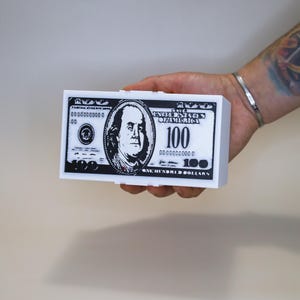 May include: A rectangular white box featuring a black and white graphic print of a one hundred dollar bill on its front surface. The design includes the portrait of Benjamin Franklin, the number 100, and the text United States of America.