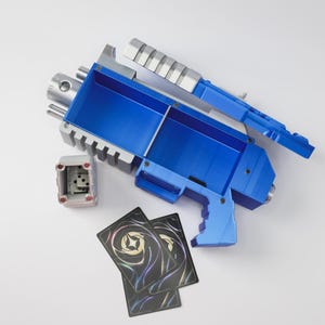 May include: A blue and silver toy gun with a box and two playing cards. The toy gun has a blue body with silver accents. The box is gray with red accents and dice inside. The playing cards have a black background with a colorful design.