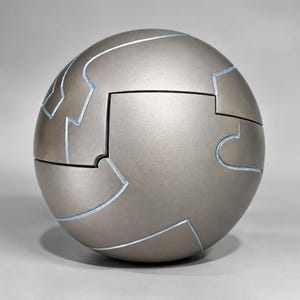 May include: A spherical puzzle object with a matte metallic gray finish. The surface is divided into interlocking segments by thin, light blue recessed lines that create a complex, puzzle-like pattern across the entire round form.