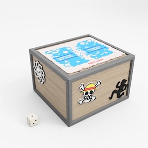 May include: A wooden box with a gray frame and a map design on top. The box features cartoon graphics of a skull with a straw hat, a silhouette of a person, and a symbol. A white die with black dots sits next to the box.