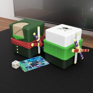 May include: Two square storage boxes, green and white, with decorative swords, a playing card, and a die. The boxes have a red band and character illustrations. The card features a character in blue and green.