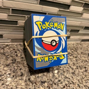 May include: A thick stack of rectangular trading cards featuring the blue and white Pokémon logo and a red and white Poké Ball graphic on the back. The cards are held together by two thin, light-colored rubber bands wrapped around the center.