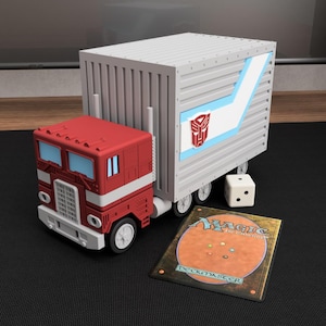 May include: A red and white toy truck with a trailer featuring a blue stripe and a red Autobot symbol. Next to the truck is a white six-sided die and a rectangular Magic The Gathering Deckmaster card with a brown and orange back design.