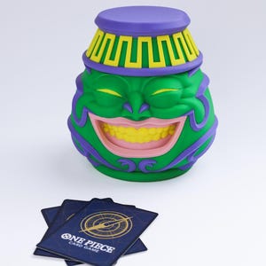 May include: A green, purple, and yellow Pot of Greed container with a smiling face, featuring a lid with a yellow geometric pattern. Beside the container are several dark blue One Piece Card Game playing cards with gold logos.