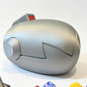 May include: A silver-colored, futuristic-looking dice box with a red accent on top. The box has a rounded, oval shape with a cylindrical detail on the side. Several colorful dice are scattered around the box.