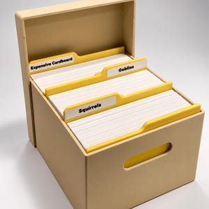 May include: A beige storage box with an open lid containing white index cards separated by yellow dividers. The dividers feature labels with the text Expensive Cardboard, Goblins, and Squirrels printed in black. The box has a cutout handle on the front.