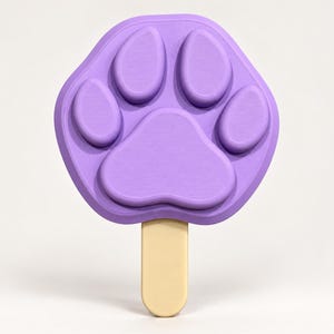 May include: A light purple paw print shaped mold with a smooth, matte finish. The mold features a large central pad and four smaller toe pads, with a light beige wooden popsicle stick inserted into the bottom edge of the design.
