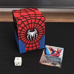 May include: A rectangular deck box featuring a red and black spiderweb pattern with a white spider emblem on the front and blue side panels, accompanied by a white six-sided die and a trading card titled Sensational Spider-Man.