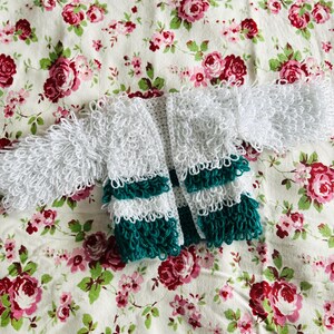 May include: A white crocheted cardigan with green stripes. The cardigan is on a floral fabric background.