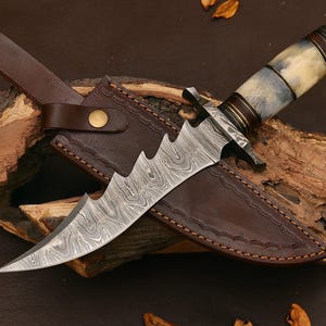 Puede incluir: Un cuchillo de acero de Damasco con borde dentado, sobre una funda de cuero marrón. El mango presenta un diseño de hueso y madera. El cuchillo y la funda se exhiben sobre una superficie de madera.