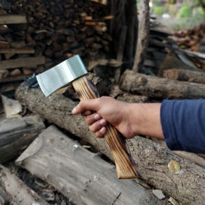 May include: A person holding a small silver axe with a wooden handle. The axe has a dark brown wooden handle with a light brown wood grain pattern. The axe is resting on a pile of logs.