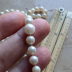 May include: A strand of luminous, round pearls held by a hand, with a second strand visible in the background. An antique ivory-colored ruler is partially visible, with measurements in inches.