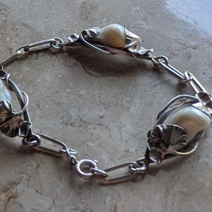 May include: Silver-toned bracelet featuring three large, off-white, oval-shaped beads. The beads are set in ornate, leaf-like settings and connected by a chain link design. The bracelet is displayed on a light-colored surface.
