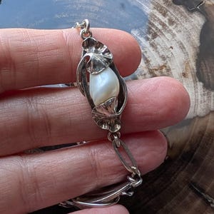 May include: A silver bracelet featuring a central pearl encased in a stylized floral design. The bracelet has a delicate chain with leaf-shaped links and a secure clasp. The pearl is white and has a smooth, lustrous surface.