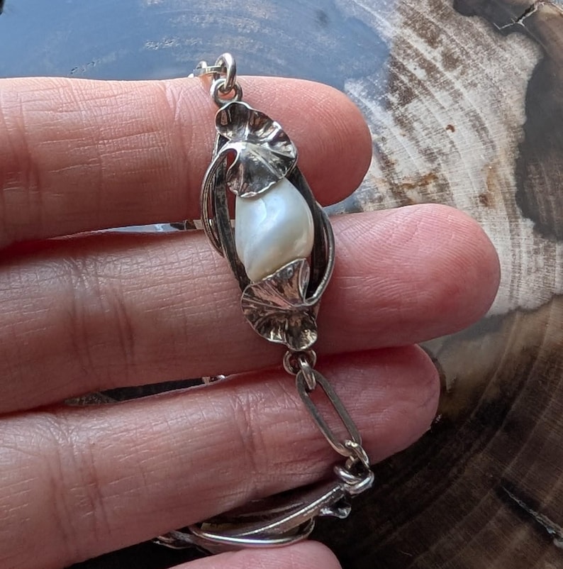 May include: A silver bracelet featuring a central pearl encased in a stylized floral design. The bracelet has a delicate chain with leaf-shaped links and a secure clasp. The pearl is white and has a smooth, lustrous surface.