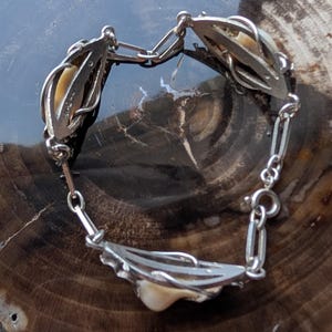 May include: A vintage silver-toned bracelet featuring ornate, openwork links. Each link holds a cream-colored, organic-shaped element. The bracelet is connected by a delicate chain and a secure clasp. The bracelet is displayed on a dark, textured surface.