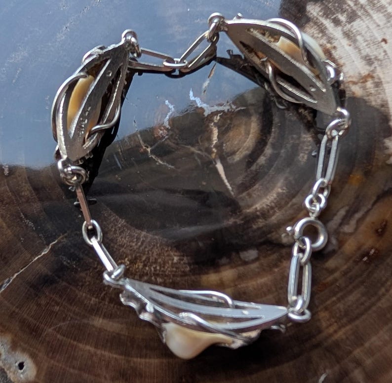 May include: A vintage silver-toned bracelet featuring ornate, openwork links. Each link holds a cream-colored, organic-shaped element. The bracelet is connected by a delicate chain and a secure clasp. The bracelet is displayed on a dark, textured surface.