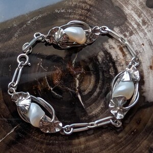 May include: A silver bracelet featuring three large, lustrous pearls set within ornate, leaf-shaped settings. The bracelet is connected by delicate links and has a secure clasp. The pearls are approximately 0.75 inches in length.