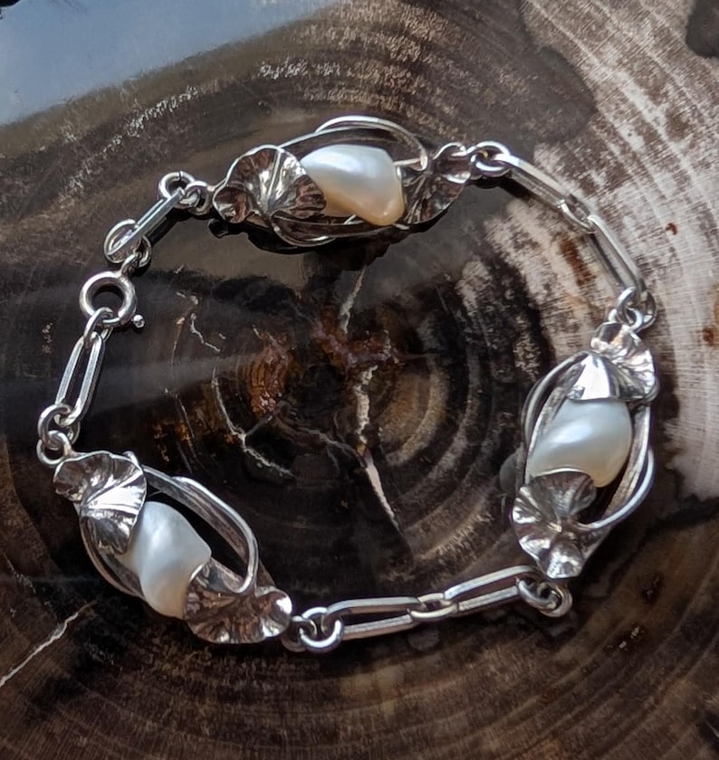 May include: A silver bracelet featuring three large, lustrous pearls set within ornate, leaf-shaped settings. The bracelet is connected by delicate links and has a secure clasp. The pearls are approximately 0.75 inches in length.