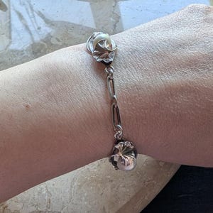 May include: Silver-toned bracelet featuring a floral design with a pearl-like bead. The bracelet has a chain link design and is worn on the wrist. The bracelet's floral elements have a detailed, textured appearance.